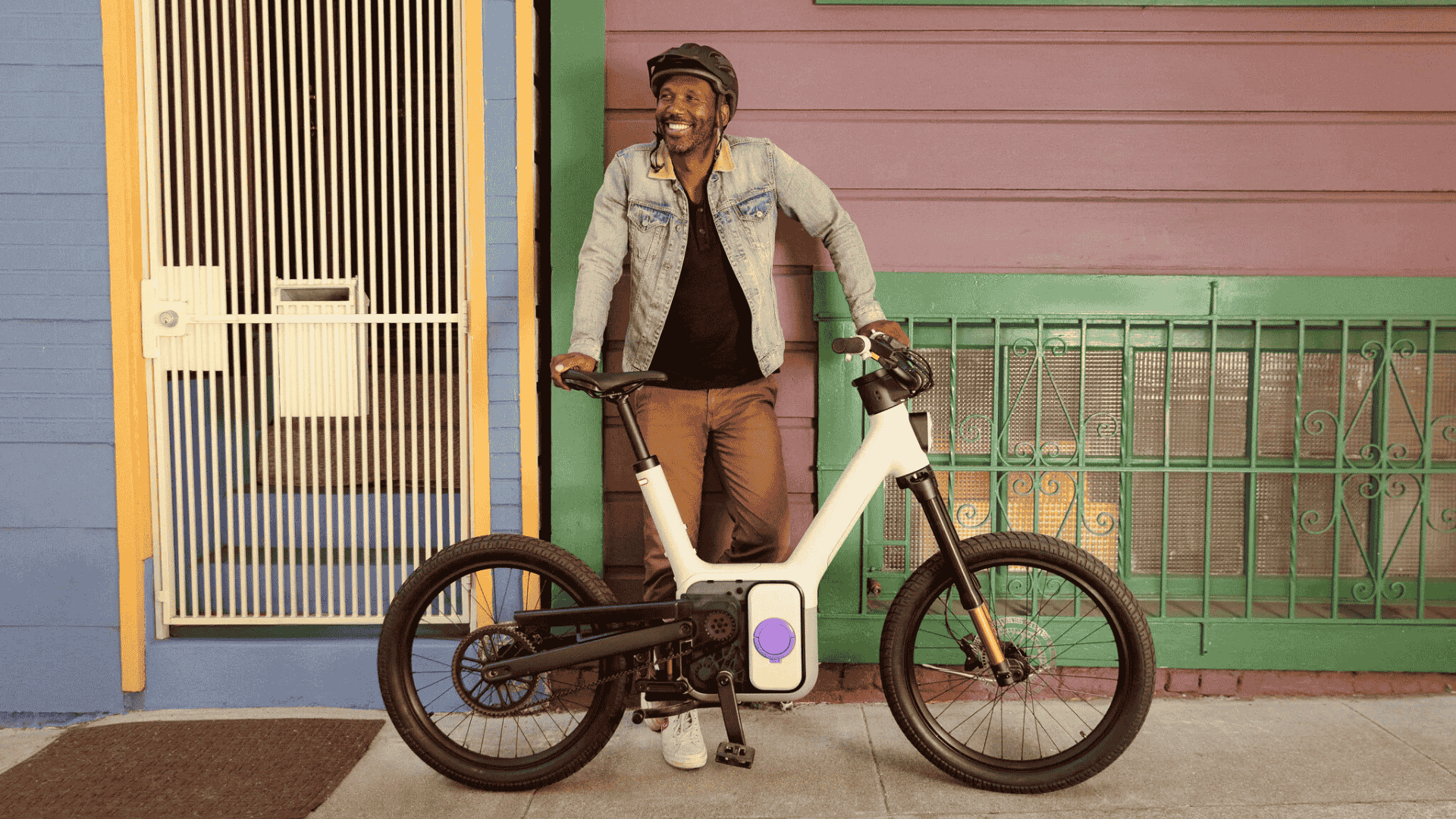Cycliste souriant posé avec l’Also TM-B de Rivian devant une façade colorée, vélo électrique urbain.