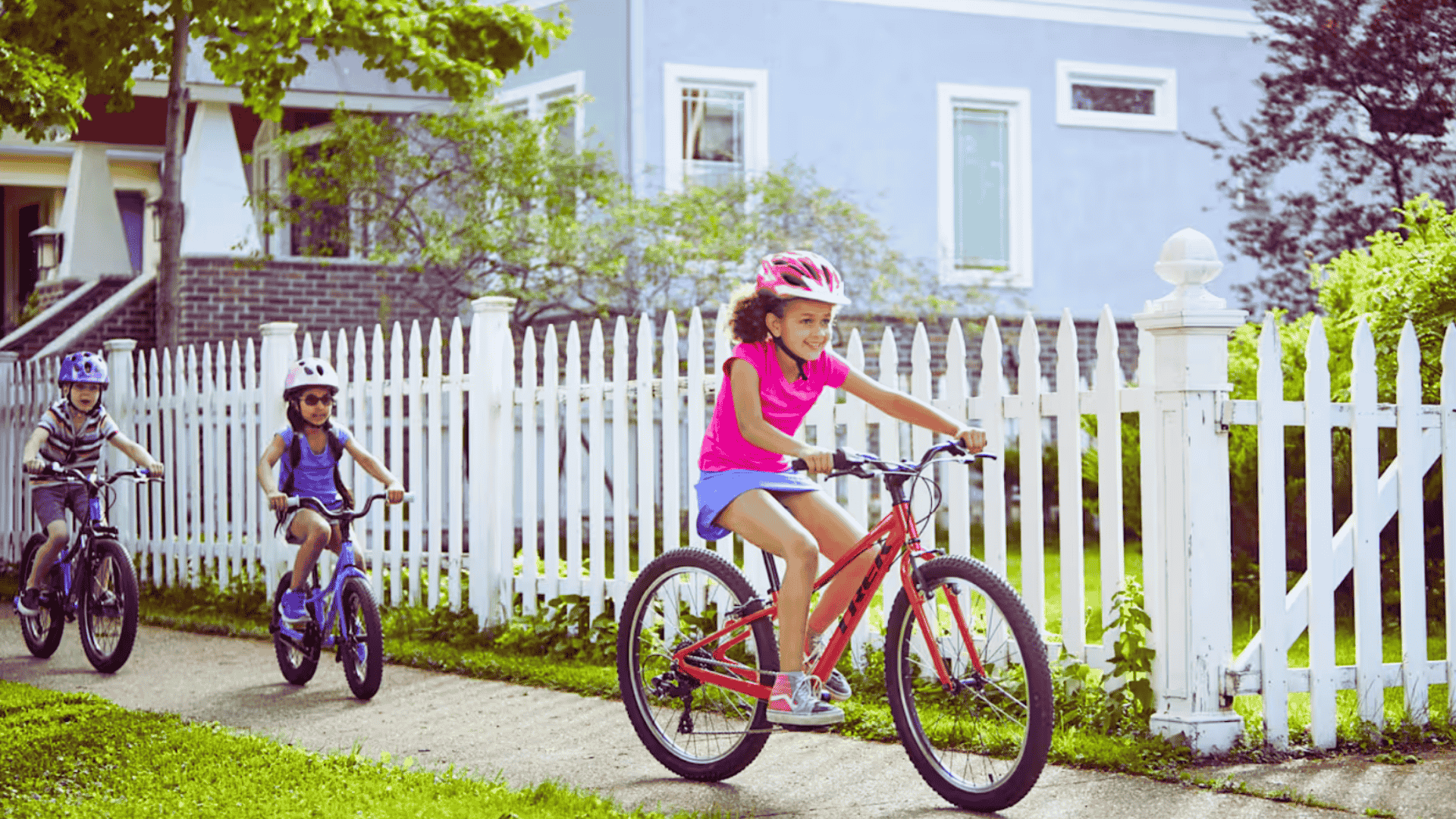 Trois enfants roulant sur des vélos Trek devant une maison, portant des casques, dans un quartier résidentiel
